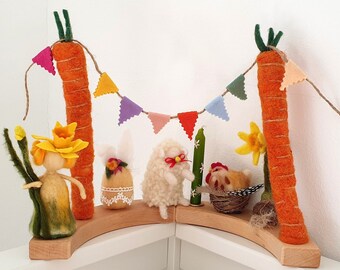 May include: A whimsical Easter display featuring felted figures and decorations. Two large orange carrot structures frame a wooden shelf holding a felted fairy, bunny, sheep, chicken, and a green candle. A colorful banner hangs between the carrots.
