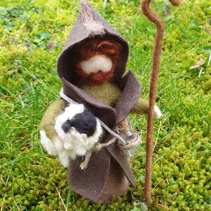 May include: A handcrafted felt figure of a wizard or shepherd, wearing a brown hooded cloak and holding a staff and a small glass bottle. The figure has a white beard and a bag. The figure is standing on green moss.