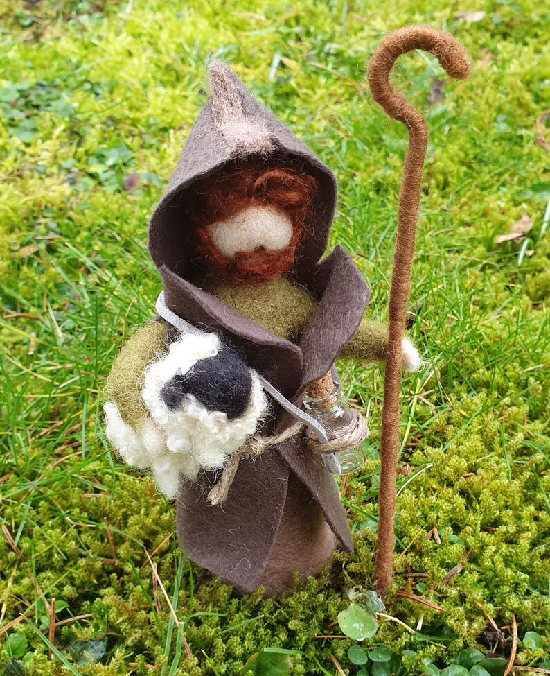 May include: A handcrafted felt figure of a wizard or shepherd, wearing a brown hooded cloak and holding a staff and a small glass bottle. The figure has a white beard and a bag. The figure is standing on green moss.