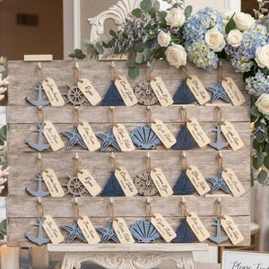 May include: A nautical-themed seating chart display. The wooden board features various sea-themed cutouts, including anchors, sailboats, starfish, and seashells, each with a name tag. The display is decorated with flowers and candles, creating an elegant ambiance.