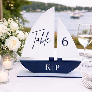 May include: A nautical-themed table number decoration shaped like a sailboat. The white sails display the word "Table" and the number "6". The navy blue hull has the initials "K | P". The decor is set on a white tablecloth with a waterfront backdrop.