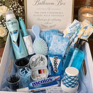 May include: A white box labelled "Wedding Bathroom Box" containing toiletries and essentials. Items include deodorant, a lint roller, hairbrush, sanitary pads, tampons, mouthwash, cups, hair ties, a sewing kit, chewing gum, mints, a Tide to go pen, and a thank you tag.