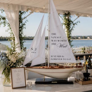 May include: A model sailboat with white fabric sails featuring the printed text 'THE VOYAGE WAS WORTH THE Wait', '11 YEARS & FOREVER TO GO', and the initials 'S J' with the date '09.18.26'. The boat has a wooden hull and sits on a black base.
