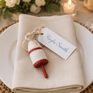 Peut inclure: Mise en place sur le thème nautique avec une assiette blanche, une serviette en lin beige et une bouée décorative. Une étiquette avec le nom "Kayla Smith" est attachée. La table comprend une fourchette et un couteau dorés et un dessous de plat tressé.