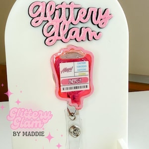May include: A pink and clear badge reel shaped like an IV bag, with the text "ENERGY" and "CAUTION CONTAINS CAFFEINE 200MG". The badge reel is displayed on a white stand with the text "Glittery Glam" in pink.