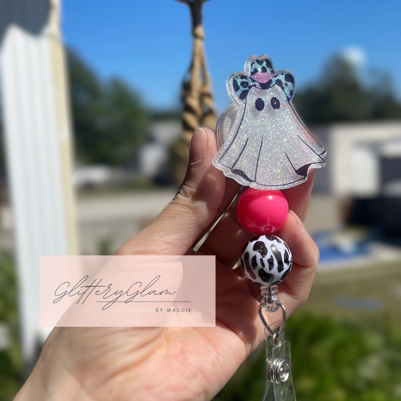 May include: A white ghost with a pink and black cow print hat, a pink bead, a black and white cow print bead, and a clear retractable badge holder. The text "Glittery Glam by Maddie" is visible in the background.