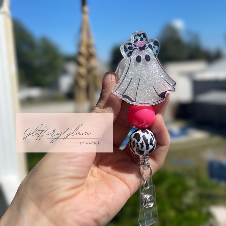 May include: A white glitter ghost wearing a pink and black leopard print cowboy hat with a pink and black leopard print bead and a pink bead on a clear retractable badge reel. The text "Glittery Glam by Maddie" is visible in the background.