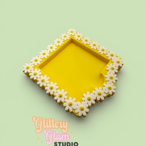 Puede incluir: Una bandeja cuadrada amarilla brillante decorada con flores de margaritas blancas alrededor de los bordes. La bandeja tiene un centro empotrado y el texto "Glittery Glam Studio" en cursiva.