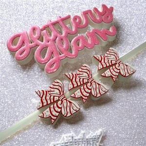May include: Pink glittery "Glittery Glam" text and three bow-shaped decorations with red and white striped patterns. The items are on a sparkly silver background, suggesting a craft or party theme.