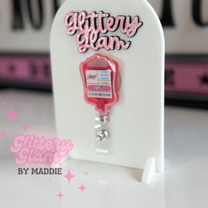 May include: A white badge holder featuring a pink glittery "Glittery Glam" logo and a red blood bag design. The badge reel has a clear plastic strap and a metal clip. The text "Glittery Glam by Maddie" is also visible.