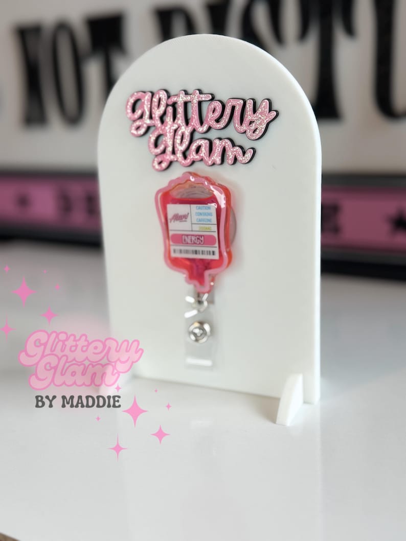 May include: A white badge holder featuring a pink glittery "Glittery Glam" logo and a red blood bag design. The badge reel has a clear plastic strap and a metal clip. The text "Glittery Glam by Maddie" is also visible.