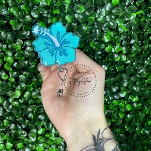 May include: A teal blue glitter hibiscus flower charm attached to a retractable badge holder. The charm is attached to a silver clip and has a clear plastic strap. The text "Glittery Glam by Maddie" is printed on a white circle.
