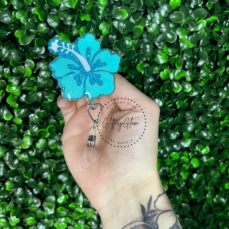 May include: A teal blue glitter hibiscus flower charm attached to a retractable badge holder. The charm is attached to a silver clip and has a clear plastic strap. The text "Glittery Glam by Maddie" is printed on a white circle.