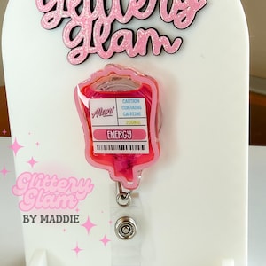 May include: A pink glitter badge reel with a blood bag design, featuring the text "Energy" and "Caution Contains Caffeine 200mg." The badge reel is attached to a white display stand with the text "Glittery Glam by Maddie."