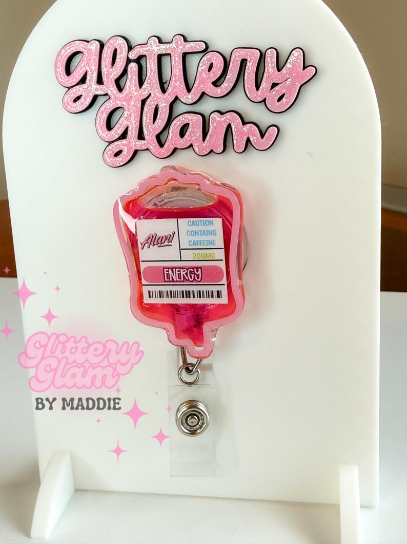 May include: A pink glitter badge reel with a blood bag design, featuring the text "Energy" and "Caution Contains Caffeine 200mg." The badge reel is attached to a white display stand with the text "Glittery Glam by Maddie."