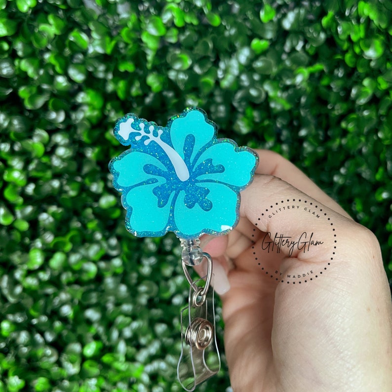 May include: A teal blue glitter hibiscus flower resin badge reel with a clear plastic retractable cord and a spring clip. The badge reel has a white glitter hibiscus flower design and the text "Glittery Glam by Maddie" in a circle.