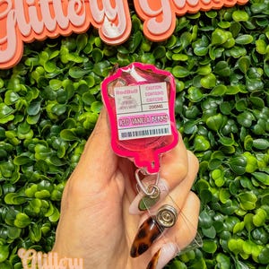 Puede incluir: Un portatarjetas rosa con forma de bolsa de goteo, con el texto "Iced Vanilla Berry" y "Red Bull The Pink Edition". El portatarjetas tiene un cord&oacute;n retr&aacute;ctil y un clip met&aacute;lico. El fondo presenta un seto verde y las palabras "Glittery Glam" en rosa.