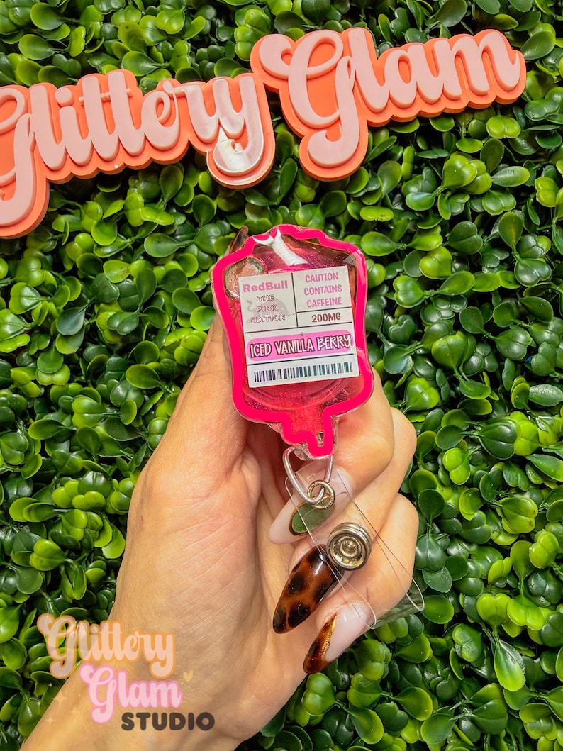 Puede incluir: Un portatarjetas rosa con forma de bolsa de goteo, con el texto "Iced Vanilla Berry" y "Red Bull The Pink Edition". El portatarjetas tiene un cord&oacute;n retr&aacute;ctil y un clip met&aacute;lico. El fondo presenta un seto verde y las palabras "Glittery Glam" en rosa.