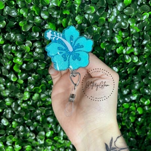 May include: A teal blue glitter hibiscus flower charm with a white center and a silver retractable badge reel. The charm is attached to a clear plastic badge holder. The charm is labeled "Glittery Glam by Maddie".