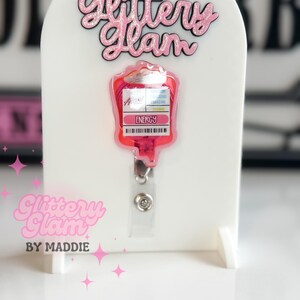 May include: A pink and red badge reel, shaped like an IV bag, with the word "Energy" and a barcode. The badge reel is displayed on a white stand, with the text "Glittery Glam" in pink script.