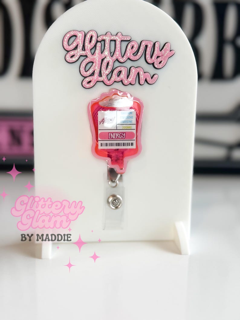 May include: A pink and red badge reel, shaped like an IV bag, with the word "Energy" and a barcode. The badge reel is displayed on a white stand, with the text "Glittery Glam" in pink script.