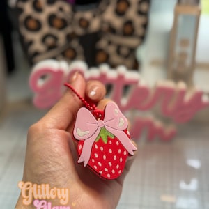Op de afbeelding: Een rode aardbei-bedel met een roze strik en groene blaadjes, bevestigd aan een rode kralenketting. De tekst "Glittery Glam Studio" is zichtbaar.
