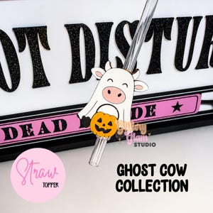 May include: A clear plastic straw featuring a decorative topper shaped like a white cow dressed as a ghost, holding a small orange jack-o'-lantern. The topper is positioned in front of a sign with black glittery text and a pink horizontal bar.