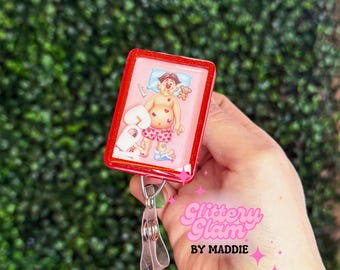 Puede incluir: Un carrete de insignia rojo con una ilustración de dibujos animados de una persona en la cama, usando pantalones cortos con estampado de corazones. El carrete de insignia tiene un clip transparente y el texto "Glittery Glam BY MADDIE" en rosa.