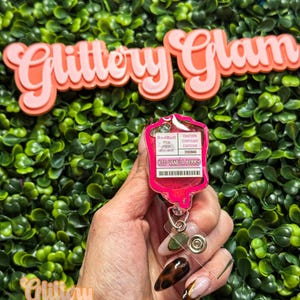 Puede incluir: Un portatarjetas rosa en forma de bolsa de goteo, con el texto "Red Bull Iced Vanilla Berry" y "Precauci&oacute;n Contiene cafe&iacute;na 200mg". El portatarjetas se muestra sobre un fondo de follaje verde, con las palabras "Glittery Glam" en rosa.