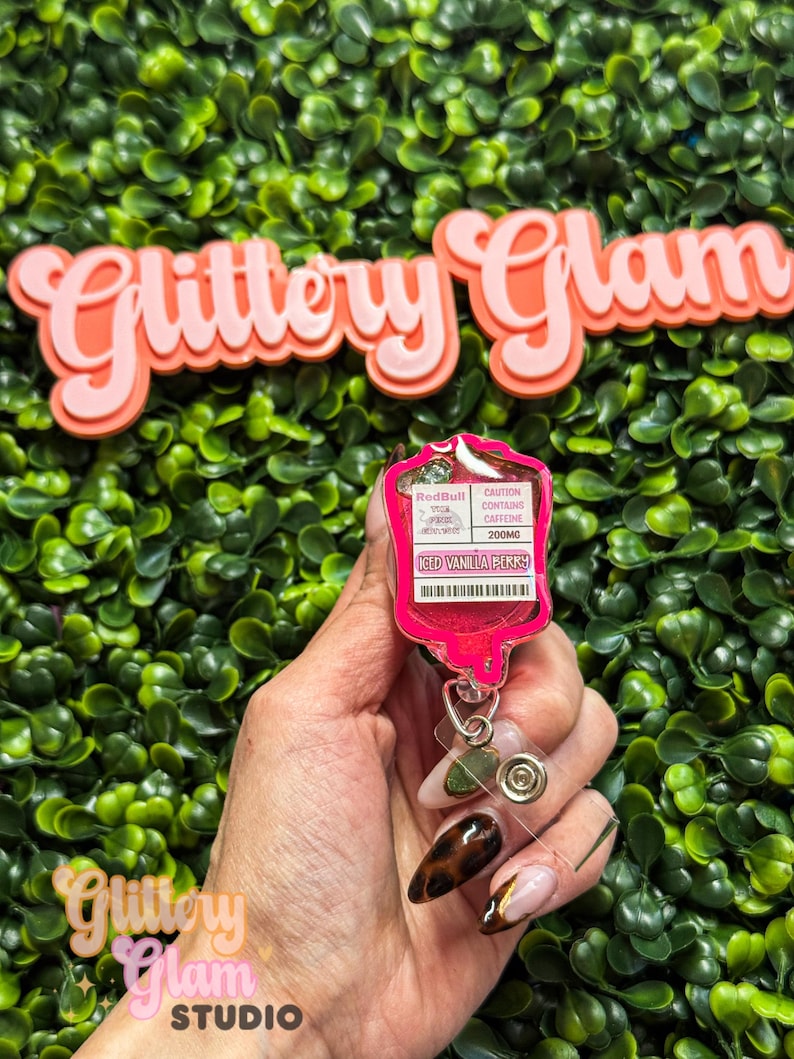 Puede incluir: Un portatarjetas rosa en forma de bolsa de goteo, con el texto "Red Bull Iced Vanilla Berry" y "Precauci&oacute;n Contiene cafe&iacute;na 200mg". El portatarjetas se muestra sobre un fondo de follaje verde, con las palabras "Glittery Glam" en rosa.