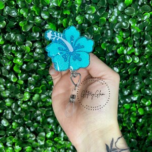 May include: A turquoise blue glitter hibiscus flower charm with a silver swivel clip. The charm is attached to a silver retractable badge reel. The reel is labeled "Glittery Glam by Maddie".