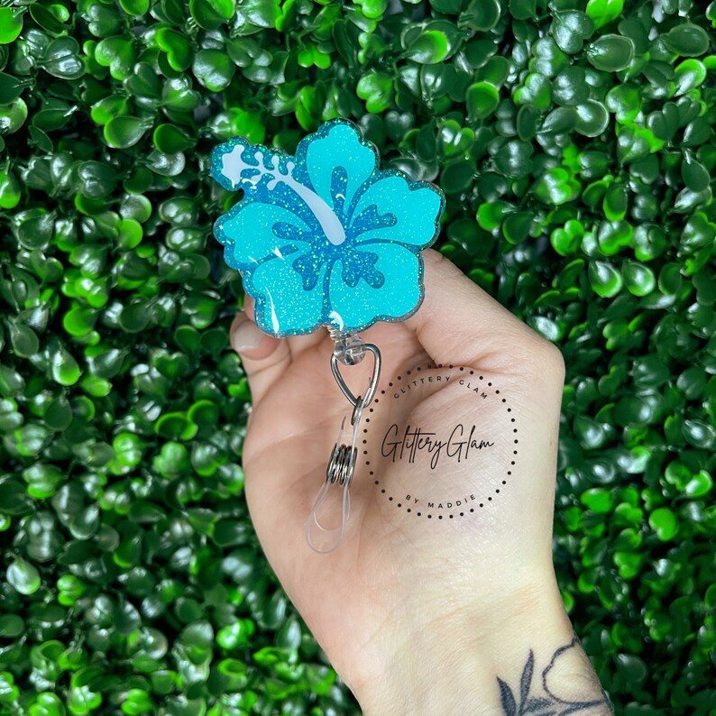 May include: A turquoise blue glitter hibiscus flower charm with a silver swivel clip. The charm is attached to a silver retractable badge reel. The reel is labeled "Glittery Glam by Maddie".