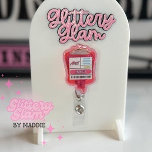 May include: A white display stand with the text "Glittery Glam" in pink glitter. A pink IV bag-shaped badge reel with the word "ENERGY" is attached. The badge reel has a clear plastic strap and a silver metal clip.
