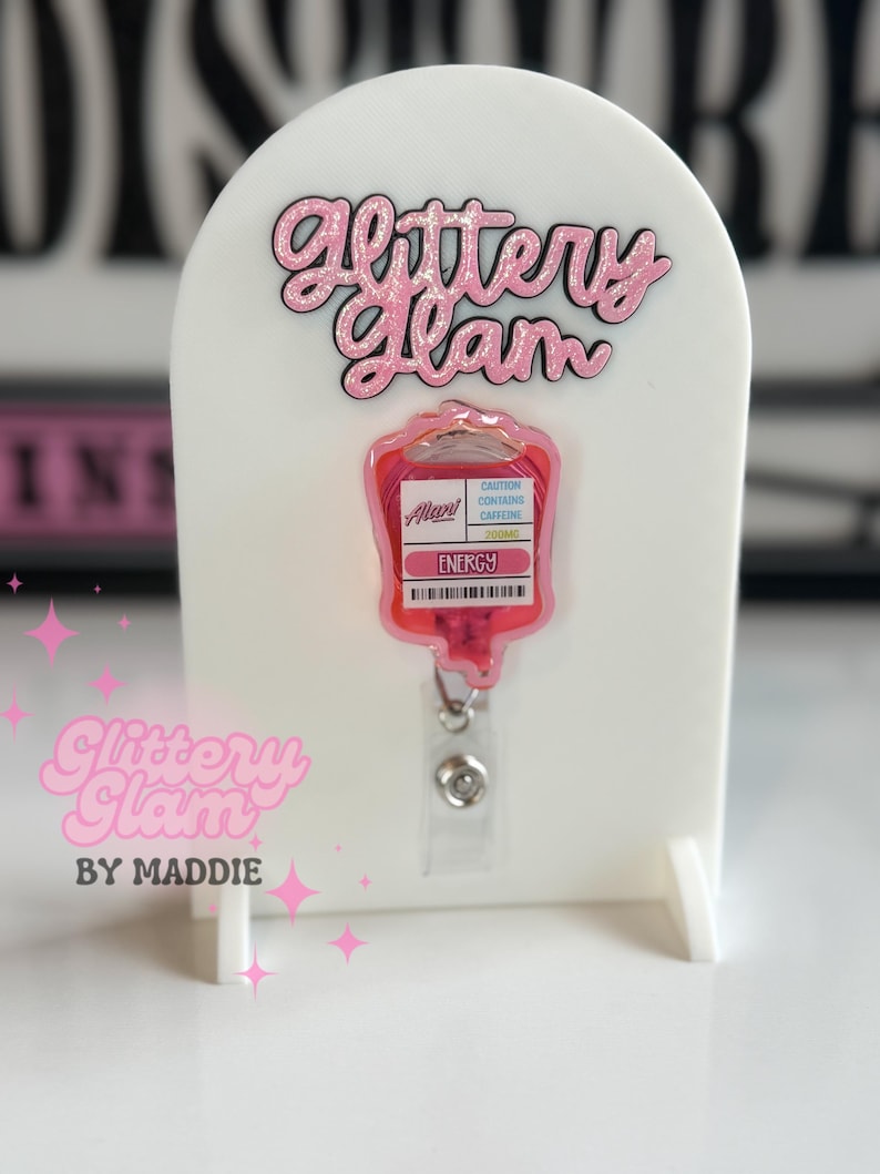 May include: A white display stand with the text "Glittery Glam" in pink glitter. A pink IV bag-shaped badge reel with the word "ENERGY" is attached. The badge reel has a clear plastic strap and a silver metal clip.