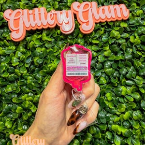 Puede incluir: Un portatarjetas rosa con un dise&ntilde;o tem&aacute;tico de Red Bull, con el texto "Iced Vanilla Perks" y "Caution Contains Caffeine 200mg". El portatarjetas se sostiene contra un fondo de follaje verde, con las palabras "Glittery Glam" en la parte superior e inferior.