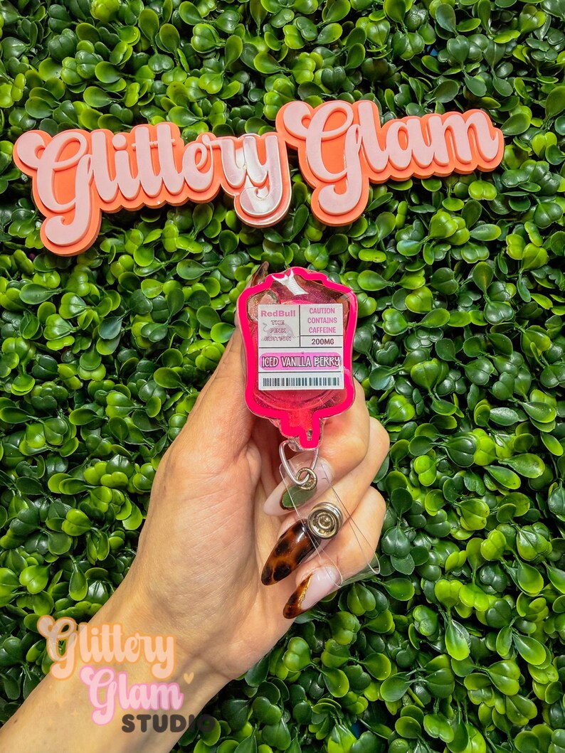 Puede incluir: Un portatarjetas rosa con un dise&ntilde;o tem&aacute;tico de Red Bull, con el texto "Iced Vanilla Perks" y "Caution Contains Caffeine 200mg". El portatarjetas se sostiene contra un fondo de follaje verde, con las palabras "Glittery Glam" en la parte superior e inferior.
