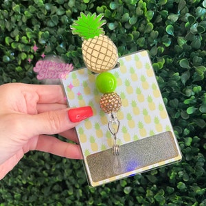 May include: A pineapple-themed badge reel with a clear plastic holder. The reel features a gold and white pineapple, a green bead, and a gold rhinestone bead. The background is a pineapple pattern.