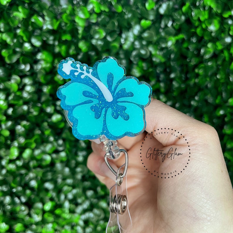May include: A turquoise blue glitter hibiscus flower with a white center, attached to a retractable badge reel. The reel is silver and has a clear plastic strap. The text "Glittery Glam by Maddie" is visible in the background.