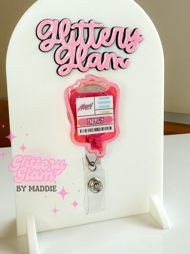 May include: A pink and clear badge reel shaped like an IV bag, with the text "ENERGY" and "CAUTION CONTAINS CAFFEINE 200MG". The badge reel is displayed on a white stand with the text "Glittery Glam" in pink.