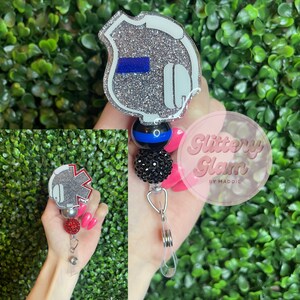 May include: Silver glitter badge holder with a blue stripe and a black and blue bead. The badge holder is attached to a silver retractable badge reel. The badge holder is on a hand with pink nail polish. The text "Glittery Glam by Maddie" is visible in the background.