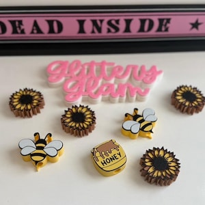 May include: A collection of decorative items featuring a bee and sunflower theme. The items include bee and sunflower-shaped embellishments, and a honey pot charm. The background includes a pink sign that reads "DEAD INSIDE" and the words "Glitter Glam".