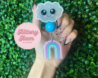 May include: A sparkly blue cloud with a smiling face, a blue bead, and a rainbow-colored retractable badge holder. The badge holder is made of clear plastic with a glitter finish. The text "Glittery Glam by Maddie" is on a pink circle.