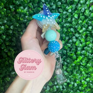 May include: A blue and white glitter starfish charm with blue and green beads attached to a clear retractable badge holder. The badge holder has a silver clip. The charm is labeled "Glittery Glam by Maddie".