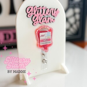 May include: A white acrylic badge holder with pink glitter text reading "Glittery Glam". A pink and red IV bag badge reel is attached. The holder also has the text "Glittery Glam by Maddie".