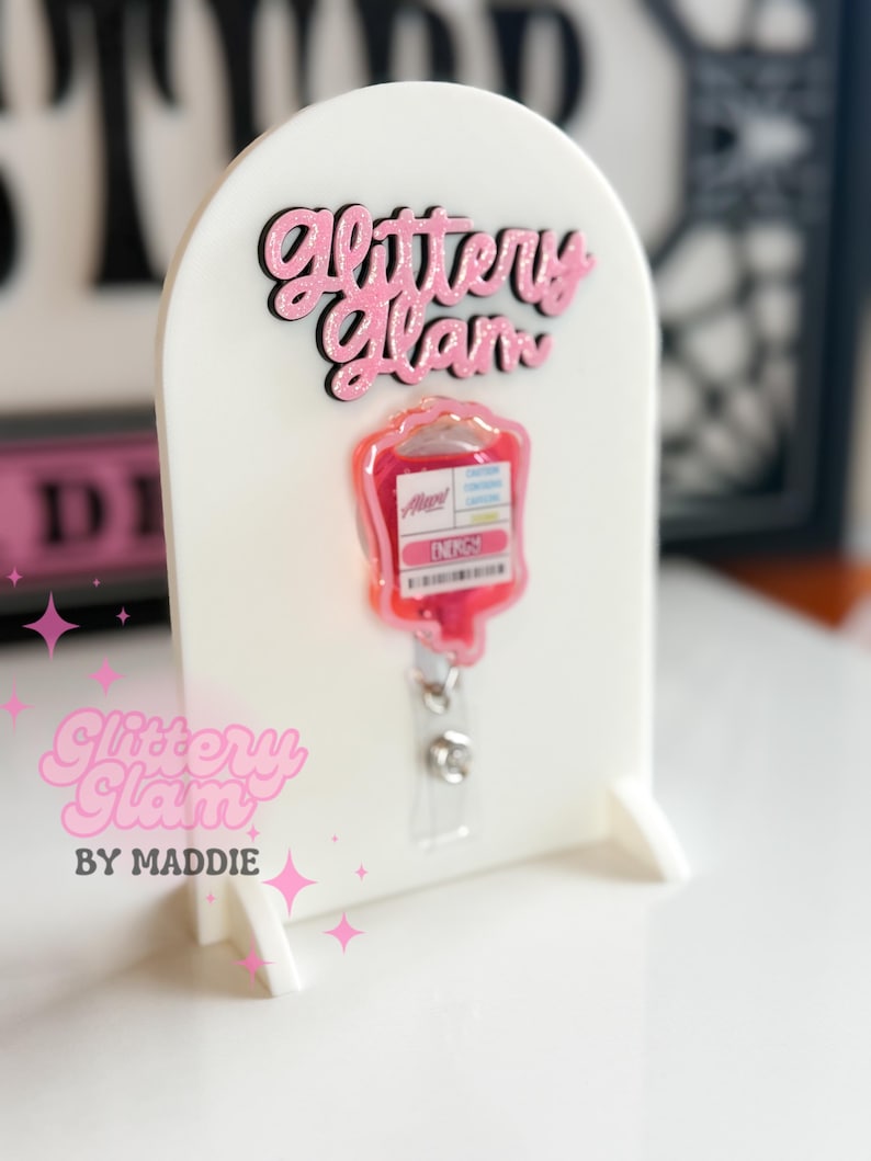 May include: A white acrylic badge holder with pink glitter text reading "Glittery Glam". A pink and red IV bag badge reel is attached. The holder also has the text "Glittery Glam by Maddie".