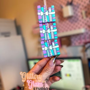 Può includere: Un ciondolo decorativo per cannucce con tre scatole regalo impilate. Ogni scatola è viola, turchese e bianca, con un fiocco bianco. Il ciondolo è tenuto da una cannuccia trasparente. Il ciondolo è di Glittery Glam Studio.