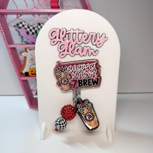 May include: A badge reel featuring a rectangular acrylic piece with pink glitter and the text "SUSPECT RUNS ON 7 BREW" alongside a smaller acrylic coffee cup charm, a red glitter bead, and a black and white checkered bead on a white display.