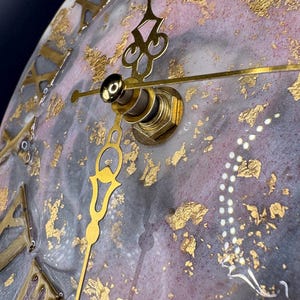 May include: Close-up of a round wall clock with a marbled gray and pink face, adorned with gold leaf accents. The clock features gold Roman numerals and ornate gold hands. The clock's mechanism is visible in the center.