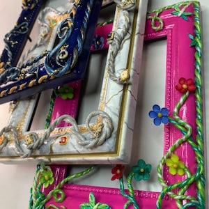 May include: Three decorative picture frames with ornate details. The top frame is navy blue with gold accents and rope-like trim. The middle frame is white with gold accents and rope-like trim. The bottom frame is pink with green vines and flowers.