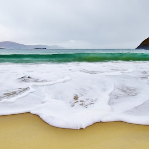 May include: A white foamy wave crashes on a sandy beach with a green ocean and mountains in the background.